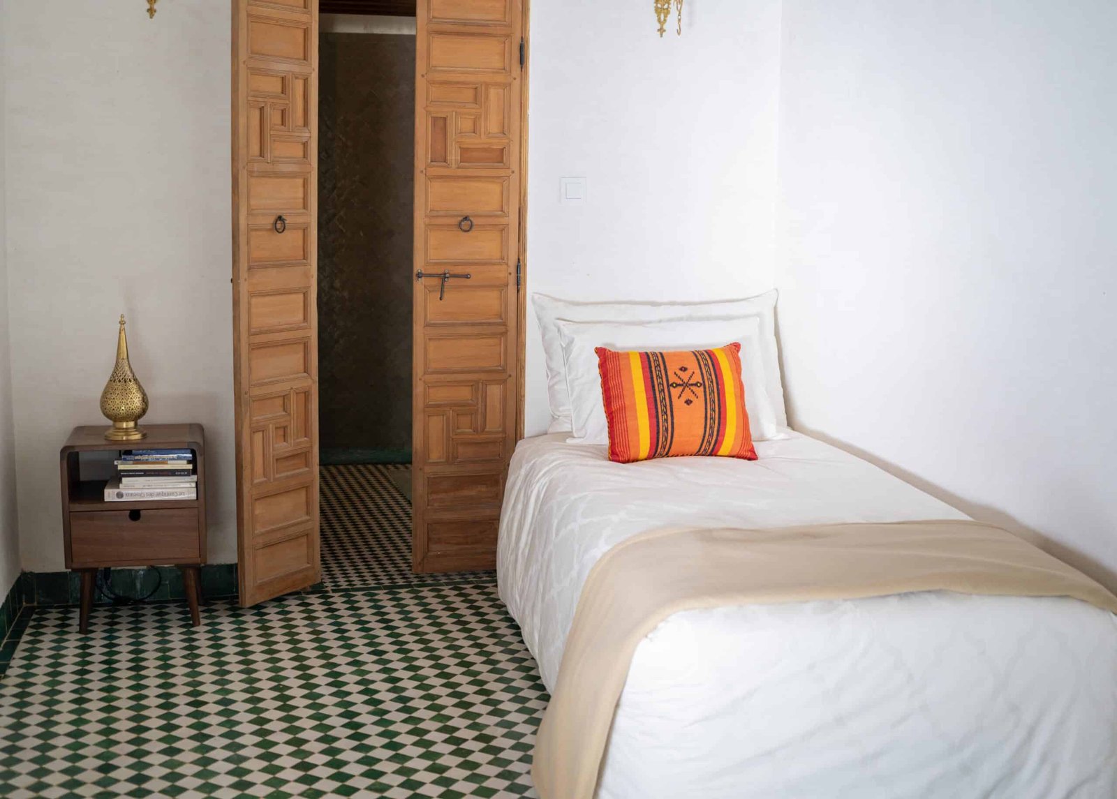 Chambre individuelle du Riad Fès avec lit simple, porte en bois sculpté et sol en zellige vert.